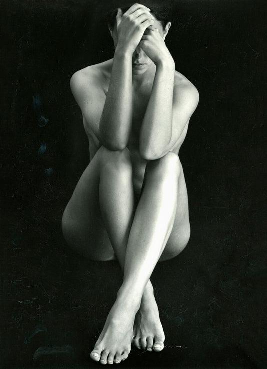Unframed black and white print ‘Classic Nude’ by Brett Weston: a nude woman with arms and legs crossed in front of her, covering her face and body. Black background.